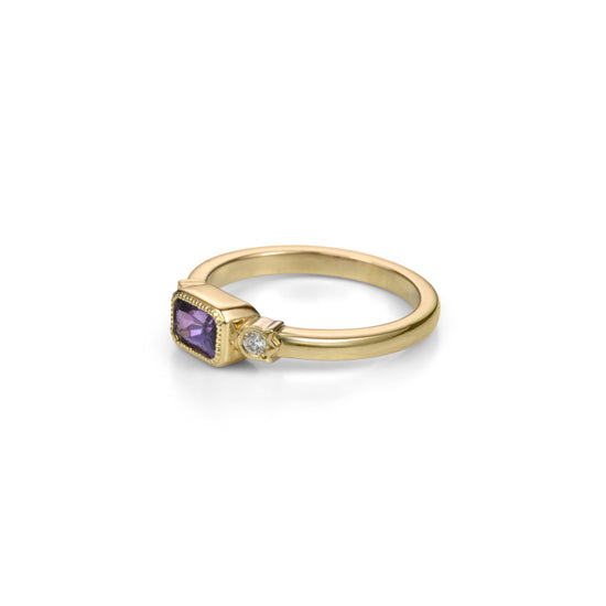 Yellow gold ring with horizontally set radiant cut royal purple sapphire in millegrained bezel and diamonds in flower shaped setting on each side in profile view.